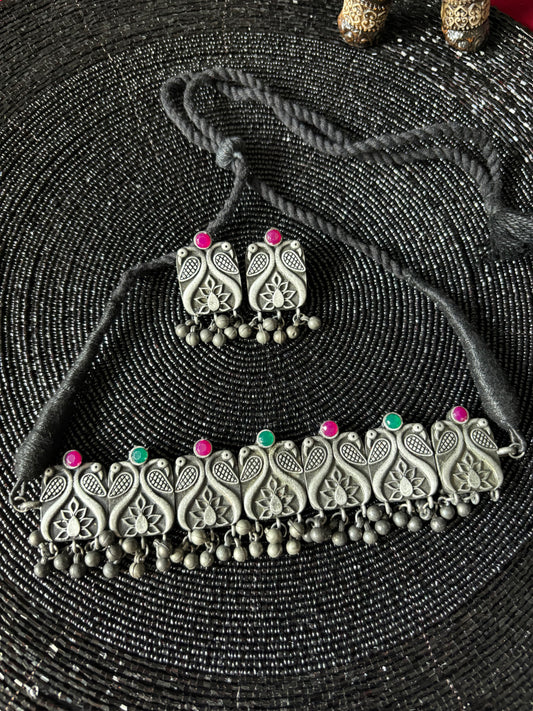 A black choker necklace with red and green beads, displayed on a textured background. The choker is adorned with intricate metalwork and bead details.