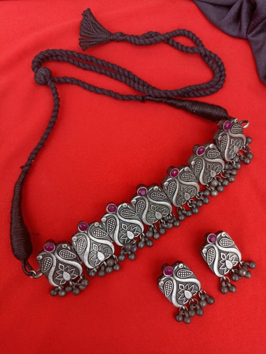 An antique style choker necklace with red beads and oxidized chain design, displayed alongside a pair of matching earrings, placed on a red background.